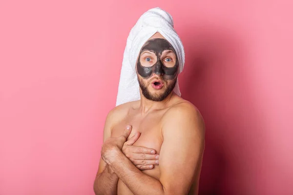 surprised young man with a cosmetic mask on his face, hiding his naked chest with his hands on a pink background.
