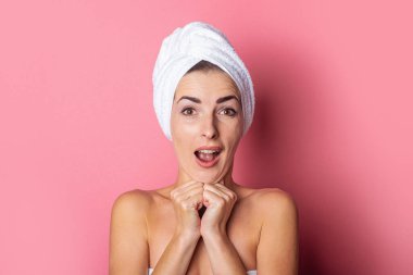 Şaşırmış genç bir kadın kafasında havluyla, çıplak omuzlarıyla ellerini çenesinin altında pembe bir arka planda tutuyor..