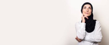 Beyaz gömlekli ve tesettürlü genç bir kadın elini çenesine dayadı ve arka planda dalgın bir yüzle yukarı baktı. Pankart.