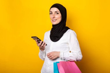 Sarı arka planda tesettürlü şirin Müslüman kadın telefon ve alışveriş torbaları tutuyor. Pankart.