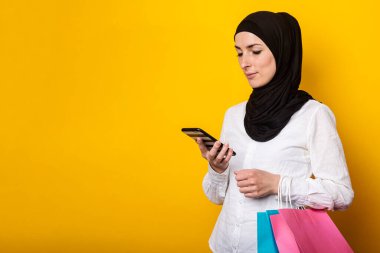 Sarışın, genç Müslüman bir kadın alışveriş çantalarını tutuyor ve sarı arka plandan sipariş vermek için telefon kullanıyor. Pankart.
