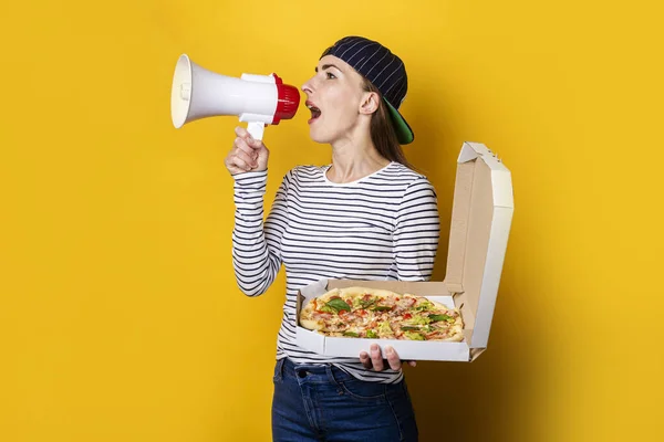 smiling young woman pizza delivery man shouting into a megaphone while holding pizza on yellow background.