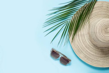 Geniş kenarlı kadın şapkası, güneş gözlüğü ve mavi arka planda bir palmiye dalı. Yaz konsepti, deniz tatili. Pankart. Düz yatış, üst görünüm.