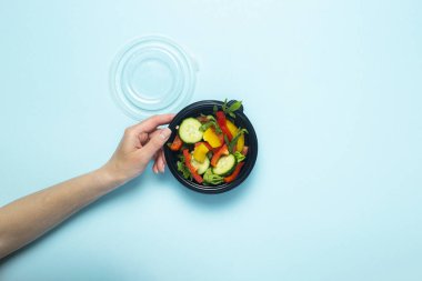 Dişi bir el, mavi arka planda sebze salatası olan tek kullanımlık plastik bir tabak tutuyor. Üst manzara, düz yatış. Pankart.