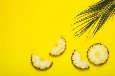 Olgun ananas dilimleri, yapraklı ananas ve sarı arka planda bir palmiye yaprağı. Üst manzara, düz uzanma.