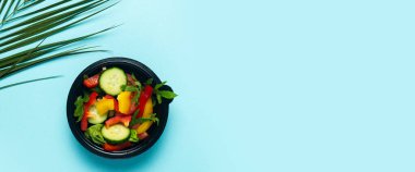 Plastik bir kasede sebze salatası ve mavi arka planda yeşil bir palmiye yaprağı. Üst manzara, düz yatış. Pankart.