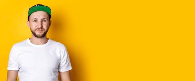 Beyzbol şapkalı, beyaz tişörtlü, sarı arka planda sakallı genç bir adam. Pankart.