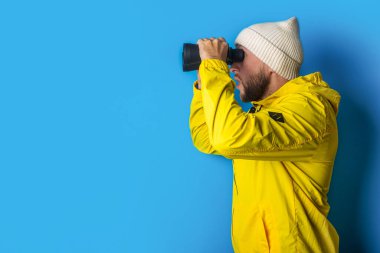 Sarı ceketli genç adam mavi arka plandaki dürbünle bakıyor..