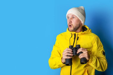 Şok olmuş sarı ceketli genç adam mavi arka plandaki dürbünle bakıyor..