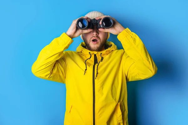 surprised young man in a yellow jacket looks through binoculars on a blue background.