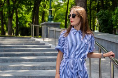 Elbiseli ve gözlüklü genç kadın parktaki merdivenlerden iniyor..