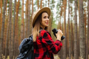 Sırt çantası, şapka ve kırmızı ekose gömlekli genç bir turist ormanda dürbün tutuyor..