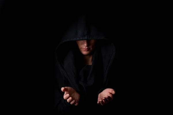 Young woman in a black hood with open palms on a dark black background.