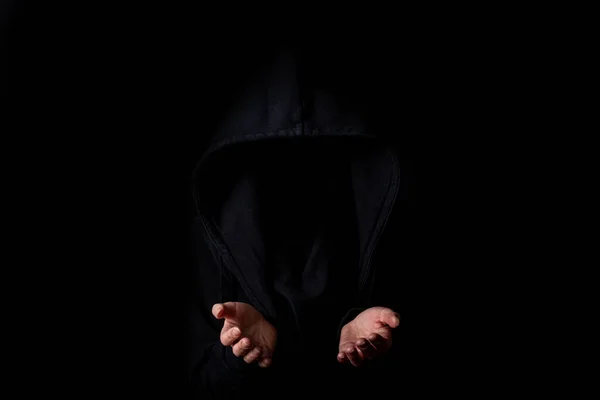 Young woman in black hood face not visible with palms outstretched against dark black background.