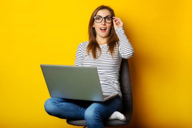 Şaşkın yüzlü genç bir kadın sandalyeye oturmuş ve dizüstü bilgisayarını izole edilmiş sarı arka planda tutuyor..
