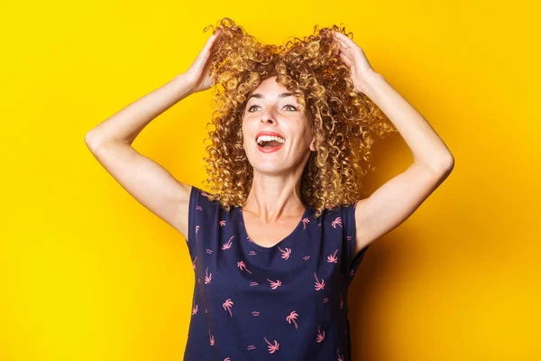 cheerful curly young woman lifts her hair up on a yellow background.