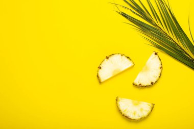 Olgun ananas dilimleri, yapraklı ananas ve sarı arka planda bir palmiye yaprağı. Üst manzara, düz uzanma.