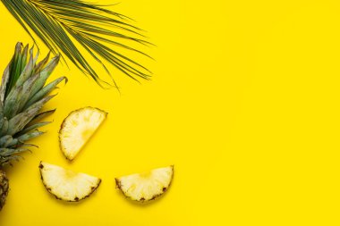 Olgun ananas dilimleri, yapraklı ananas ve sarı arka planda bir palmiye yaprağı. Üst manzara, düz uzanma.