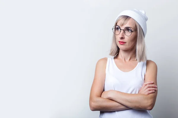 Serious young woman holding her arms crossed in a white T-shirt on a light background.