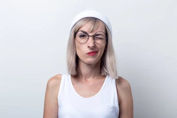 Blond young woman winking in glasses, a white hat on a light background.
