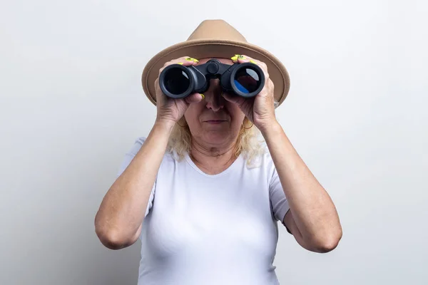 Old woman in a hat looking through binoculars on a light background