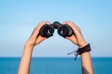 Dişi eller, gökyüzü ve denizin arka planına karşı dürbünü tutarlar..