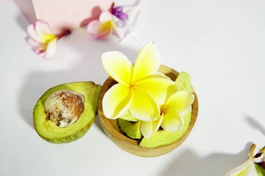 Beyaz arka planda sarı frangipani çiçekleri olan ahşap bir kasede taze avokado yarımları ve dilimler tropikal, sağlıklı ve minimalist bir kompozisyon oluşturur..