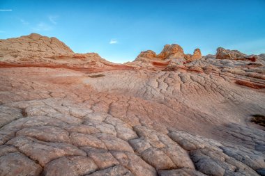 Arka planda mavi gökyüzü olan kayalık bir çöl manzarası. Gökyüzü açık ve güneş parlıyor.