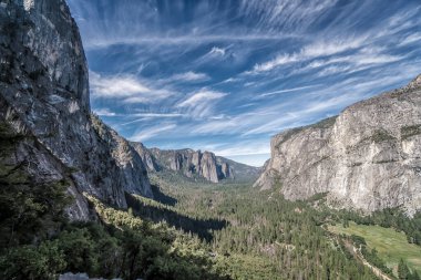 Aralarında vadi olan bir dağ sırası. Gökyüzü bazı bulutlarla mavidir. Vadi ağaçlar ve otlarla doludur.