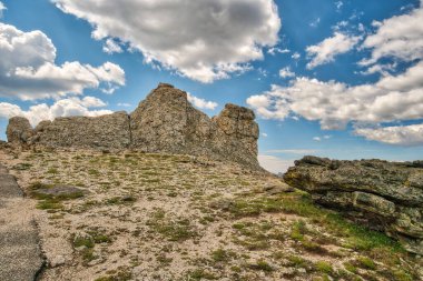 Önünde büyük bir kaya olan kayalık bir yamaç. Gökyüzü mavi ve bulutlu.