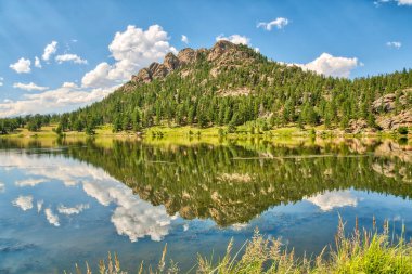 Bir dağ sırası bir gölün suyuna yansıtılır. Gökyüzü açık ve mavi, ağaçlar gür ve yeşildir. Sahne huzurlu ve sakin, su ve dağlarla güzel bir manzara yaratıyor.