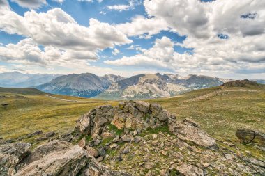 Arka planda dağ olan kayalık bir alan. Gökyüzü bulutlu ve güneş parlıyor