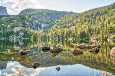 Ağaçlar ve kayalarla çevrili güzel bir göl. Su sakin ve durgun, yukarıdaki ağaçları ve gökyüzünü yansıtıyor. Huzurlu ve huzurlu bir sahne.