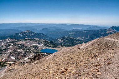 Önünde göl olan bir dağ sırası. Gökyüzü açık ve mavi. Dağlar ağaçlarla kaplı ve göl kayalarla çevrili.