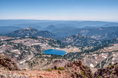 Önünde göl olan güzel bir dağ manzarası. Göl ağaçlar ve kayalarla çevrilidir ve gökyüzü açık ve mavidir. Sahne huzurlu ve huzurlu. Rahatlamak için harika bir yer.