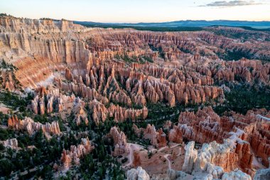 Birçok kırmızı kaya oluşumuna sahip bir kanyonun panoramik görüntüsü. Gökyüzü turuncu ve mavi renklerin karışımıdır.