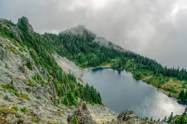 Önünde göl olan bir dağ. Göl ağaçlarla çevrili ve gökyüzü bulutlu.