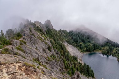 Önünde göl, arkasında sisli bir gökyüzü olan bir dağ. Göl ağaçlarla çevrilidir ve dağ kayalarla kaplıdır. Huzurlu ve huzurlu bir sahne.