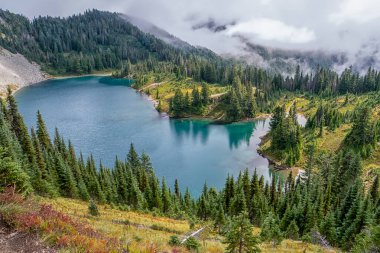 Ağaçlar ve dağlarla çevrili güzel bir göl. Göl mavi ve sakindir, ağaçlar yeşil ve yemyeşildir. Sahne huzurlu ve huzurlu. Huzur ve rahatlama hissi uyandırıyor.