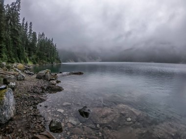 Arkasında bulutlu bir gökyüzü olan bir göl. Su sakin ve berrak. Gölün etrafındaki ağaçlar uzun ve yeşildir.