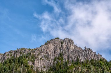 Arkasında bulutlu bir gökyüzü olan bir dağ. Dağ ağaçlar ve kayalarla kaplı. Gökyüzü mavi ve bulutlu.