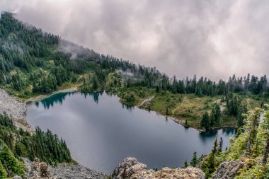 Ağaçlar ve dağlarla çevrili bir göl. Gökyüzü bulutlu ve göl sakin. Huzurlu ve huzurlu bir sahne.