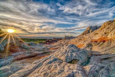 Üzerinde güneş parlayan kayalık bir manzara. Güneş batıyor ve gökyüzü bulutlu.