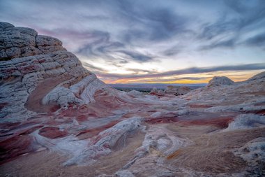 Arkasında güzel bir günbatımı olan kayalık, çorak bir manzara. Gökyüzü bulutlarla dolu, karamsar bir atmosfer yaratıyor.