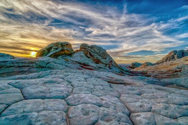 Arkasında güzel bir günbatımı olan kayalık bir manzara. Gökyüzü bulutlarla dolu, karamsar bir atmosfer yaratıyor.