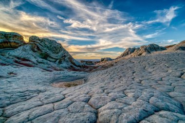 Ortasında küçük bir gölet olan kayalık, çorak bir arazi. Gökyüzü bulutlu ve güneş batıyor.