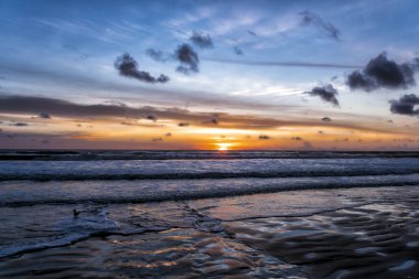 Gökyüzü güzel bir mavi ve pembemsi bir tonu var. Güneş okyanusun üzerinde batıyor, suyun üzerine sıcak bir parıltı saçıyor. Sahne huzurlu ve sakin, dalgalar yavaşça kıyıya vuruyor.