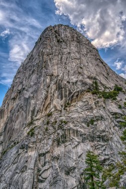 Büyük bir zirvesi ve bulutlu bir gökyüzü olan bir dağ. Dağ çok uzun ve kayalıktır. Gökyüzü bulutlu ve güneş bulutların arasında parlıyor.