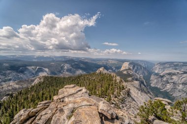 Arka planda açık mavi gökyüzü olan bir dağ sırası. Dağlar ağaçlarla örtülmüş, gökyüzü bulutlarla doludur.