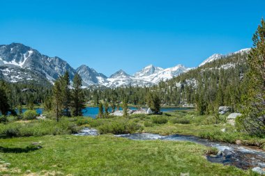 İçinden nehir akan güzel bir dağ manzarası. Gökyüzü açık ve mavi, ağaçlar gür ve yeşildir. Huzurlu ve huzurlu bir sahne, sakin ve sakin.